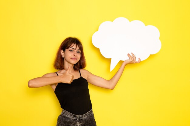 Front view of young female in black shirt and grey jeans holding huge white sign on yellow wall