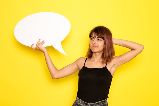 Front view of young female in black shirt and grey jeans holding white sign on yellow wall