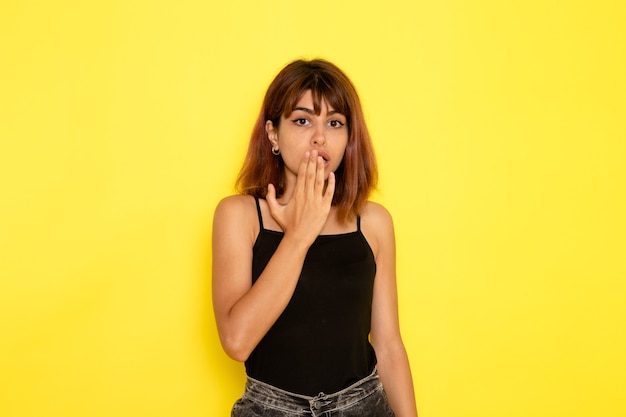 Foto gratuita vista frontale della giovane donna in camicia nera e jeans grigi in posa sulla parete gialla