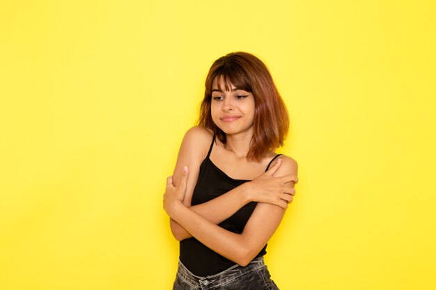 Front view of young female in black shirt and grey jeans shivering on the yellow wall