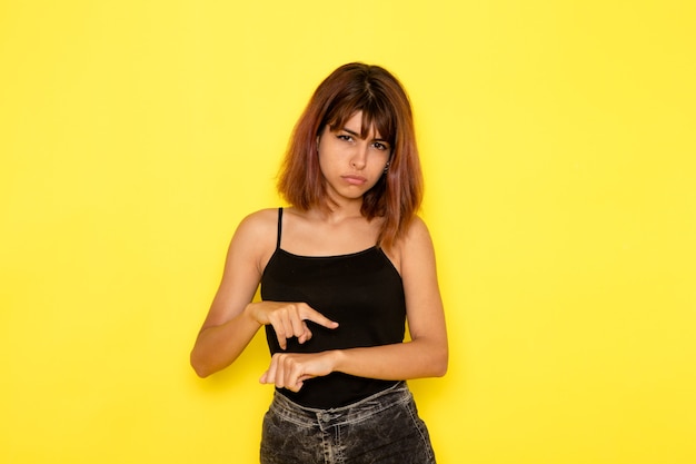 Foto gratuita vista frontale della giovane donna in camicia nera e jeans grigi che mostra il suo polso sulla parete gialla