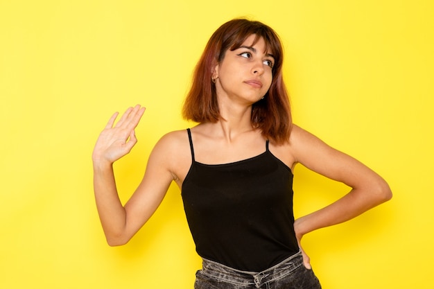 Front view of young female in black shirt and grey jeans simply posing on yellow wall