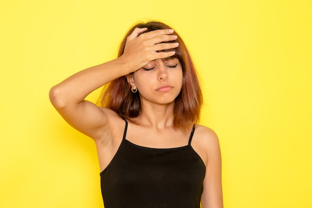 Front view of young female in black shirt and grey jeans suffering from headache on yellow wall