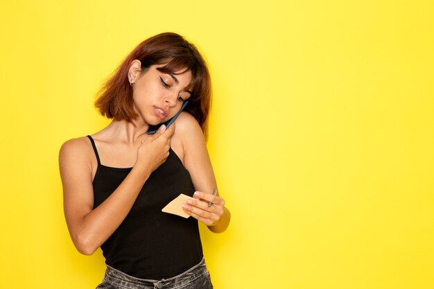 Front view of young female in black shirt and grey jeans talking on the phone on yellow wall