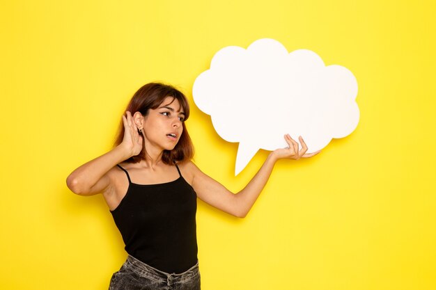 Front view of young female in black shirt holding a big white sign on light-yellow wall