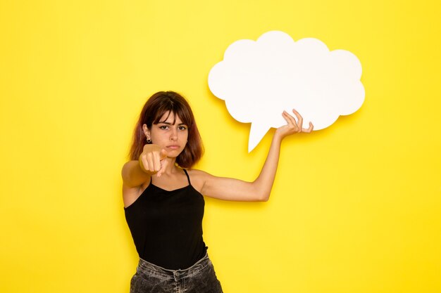 Front view of young female in black shirt holding big white sign on light-yellow wall