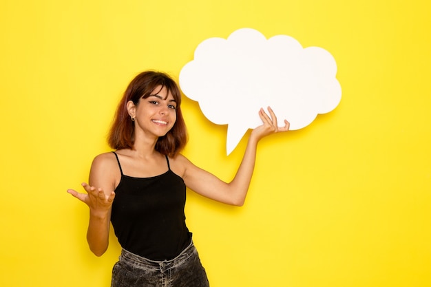 Front view of young female in black shirt holding a big white sign and smiling on light-yellow wall