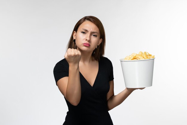 Вид спереди молодая женщина в черной рубашке держит картофельные чипсы, показывая кулак на белой поверхности