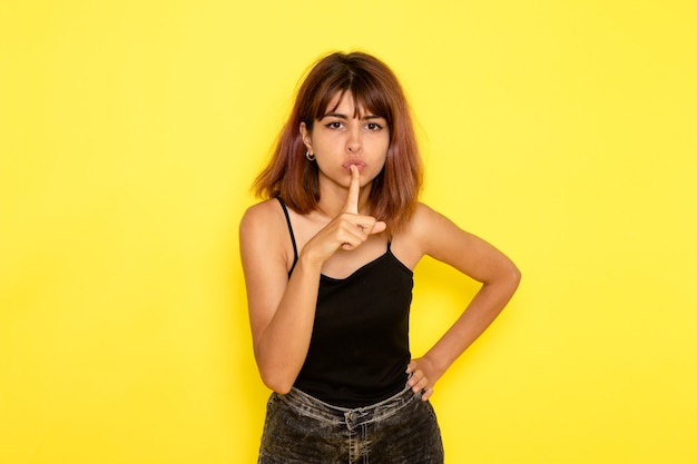 Foto gratuita vista frontale della giovane donna in camicia nera in posa e che mostra il segno di silenzio sul muro giallo chiaro