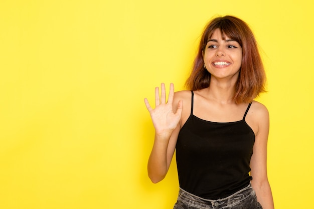 Front view of young female in black shirt waving her hand on light yellow wall