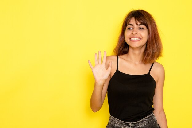 Front view of young female in black shirt waving her hand on light yellow wall