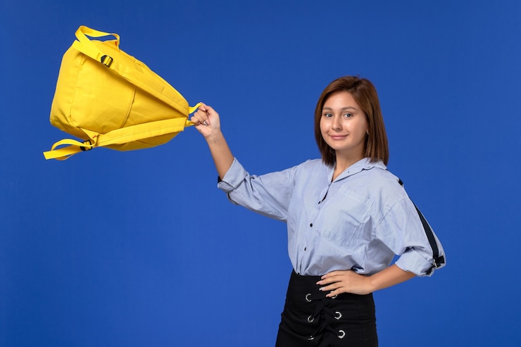 मुफ़्त फोटो front view of young female in blue shirt black skirt holding her yellow backpack with smile on the light-blue wall