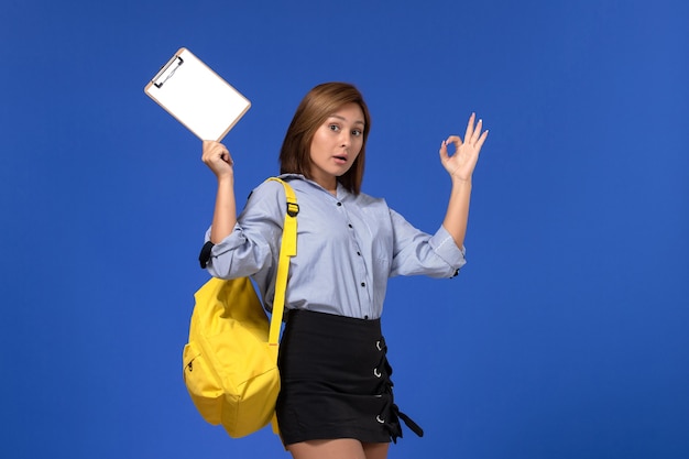 水色の壁にポーズをとってメモ帳を保持している黄色のバックパックを身に着けている青いシャツの黒いスカートの若い女性の正面図