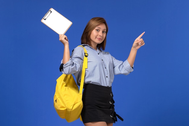 Front view of young female in blue shirt black skirt wearing yellow backpack holding notepad smiling on light blue wall