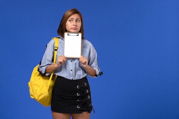 Front view of young female in blue shirt black skirt wearing yellow backpack holding notepad thinking on light blue wall