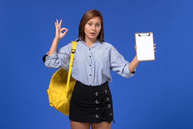 Вид спереди молодой женщины в синей рубашке с черной юбкой в желтом рюкзаке и с блокнотом