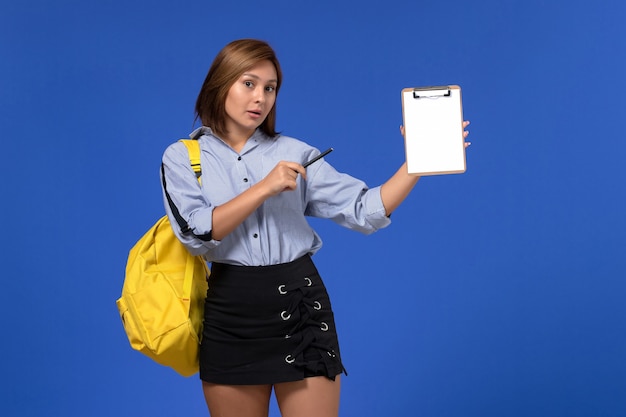 Front view of young female in blue shirt black skirt wearing yellow backpack and holding pen with notepad on blue wall