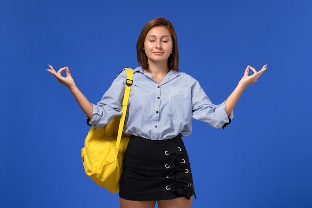 水色の壁で瞑想する黄色のバックパックを身に着けている青いシャツの黒いスカートの若い女性の正面図
