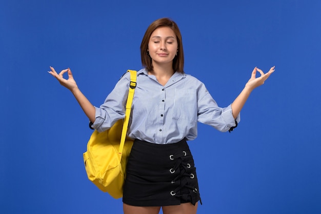 Front view of young female in blue shirt black skirt wearing yellow backpack meditating on the light-blue wall