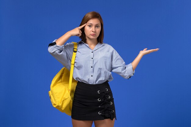 Front view of young female in blue shirt black skirt wearing yellow backpack posing on light blue wall