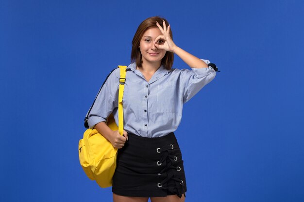 水色の壁に笑みを浮かべてポーズをとって黄色のバックパックを身に着けている青いシャツの黒いスカートの若い女性の正面図