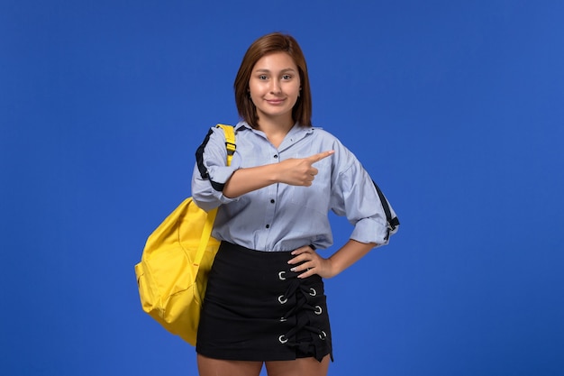 Front view of young female in blue shirt black skirt wearing yellow backpack posing and smiling on the light-blue wall