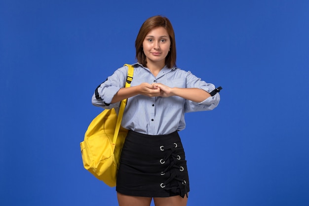 Front view of young female in blue shirt black skirt wearing yellow backpack posing smiling on the light-blue wall