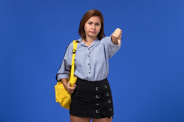 Front view of young female in blue shirt black skirt wearing yellow backpack showing unlike sign on light blue wall