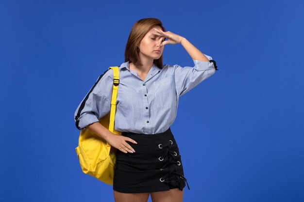 Вид спереди молодой женщины в синей рубашке с черной юбкой в желтом рюкзаке, закрывающей нос на голубой стене