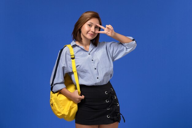 Front view of young female in blue shirt black skirt wearing yellow backpack smiling and posing on light blue wall