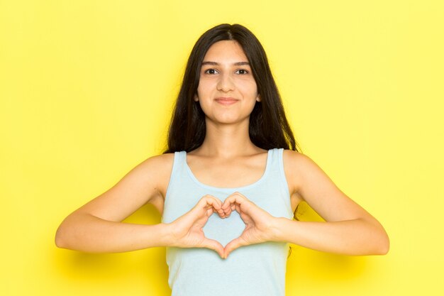 A front view young female in blue shirt posing and showing heart sign on the yellow background girl pose model beauty young