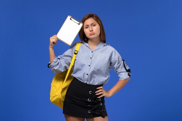 Front view of young female in blue shirt wearing yellow backpack holding notepad thinking on light blue wall