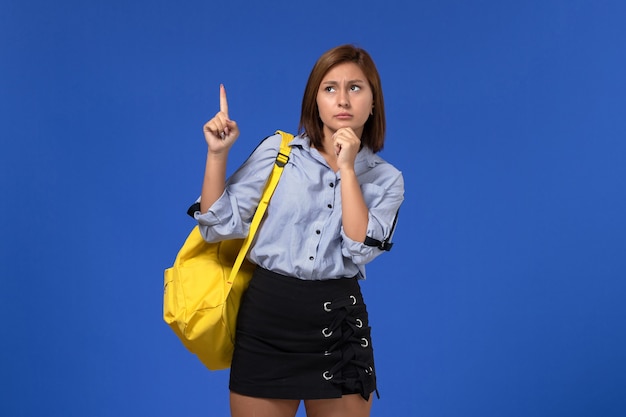 Вид спереди молодой женщины в синей рубашке в желтом рюкзаке позирует и думает на синей стене