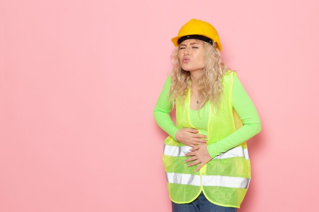 Front view young female builder in green construction suit helmet having a stomachache on the pink space architecture construction job work lady