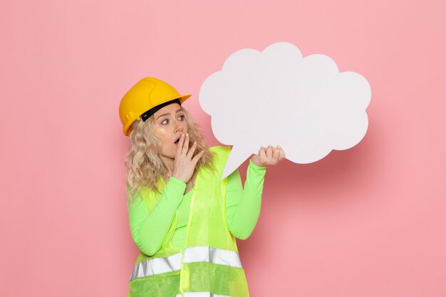 Front view young female builder in green construction suit helmet holding a big white sign on the pink space architecture construction work job