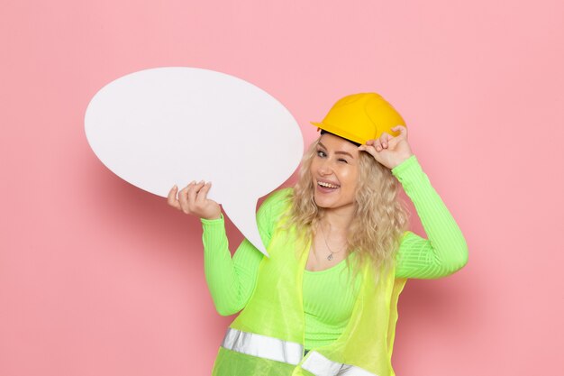 Front view young female builder in green construction suit helmet holding a big white sign while winking on the pink space architecture construction work