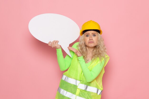 Front view young female builder in green construction suit helmet holding white sign on the pink space  job