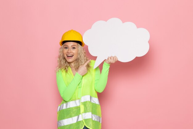 Front view young female builder in green construction suit helmet holding white sign with happy expression on the pink space 