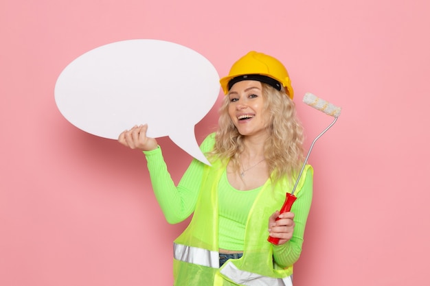 Front view young female builder in green construction suit helmet just posing with white sign smiling on the pink space architecture construction job work lady