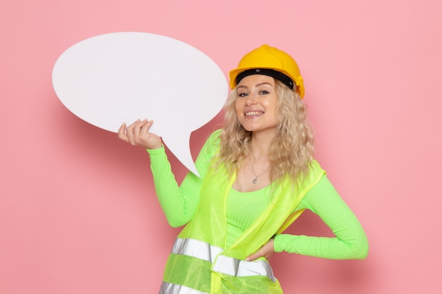 Вид спереди молодая женщина-строитель в зеленом строительном костюме в желтом шлеме с большим белым знаком, улыбаясь в розовом пространстве, работа, архитектура, строительство