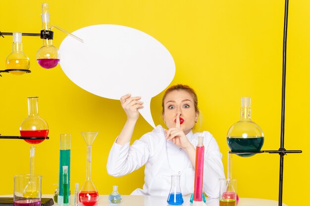 Front view young female chemist sitting in white suit with different solutions holding white sign on the yellow floor chemistry science  process