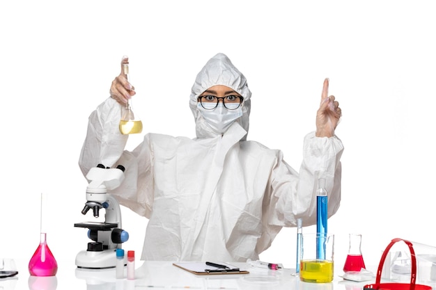 Вид спереди молодая женщина-химик в специальном защитном костюме, держащая фляжку на белом столе, лаборатория химического вируса covid