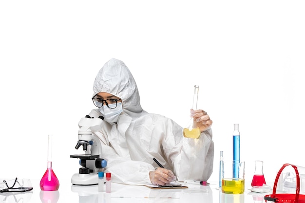 Вид спереди молодая женщина-химик в специальном защитном костюме, держащая колбу с раствором и использующая микроскоп на белом фоне, лабораторный вирус химии ковид