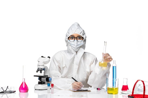 Front view young female chemist in special protective suit holding flask with solution and writing on white background lab covid chemistry virus