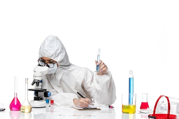 Front view young female chemist in special protective suit holding tiny flask using microscope on white background lab covid chemistry virus