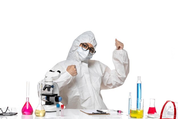 Вид спереди молодая женщина-химик в специальном защитном костюме, сидящая перед столом с растворами на светлом белом фоне, вирусная лаборатория, химия здоровья covid