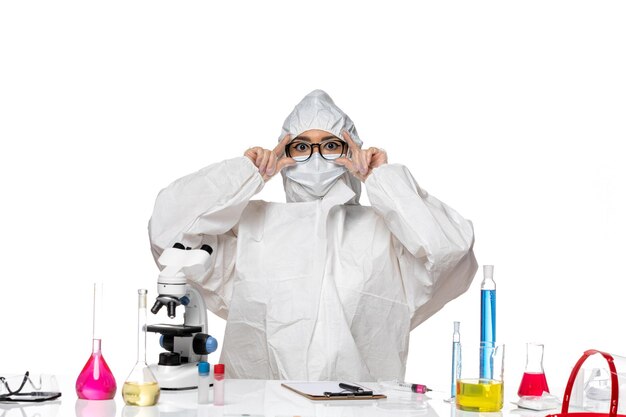 Front view young female chemist in special protective suit sitting in front of table with solutions on white desk virus lab covid health chemistry