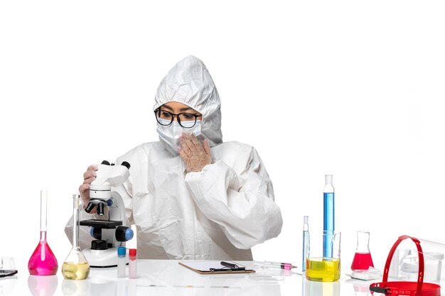 Front view young female chemist in special protective suit using microscope on light white background virus lab covid health chemistry