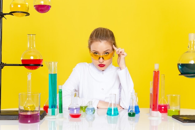 Free photo front view young female chemist in white suit in front of table with ed solutions working with them mixing up on the yellow space chemistry science   job