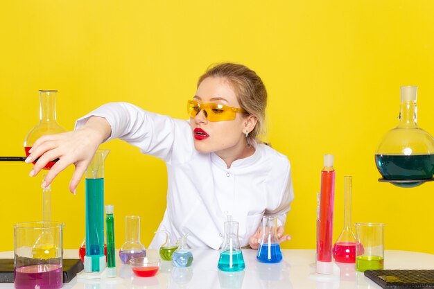 Front view young female chemist in white suit in front of table with ed solutions working with them on the yellow space chemistry  job